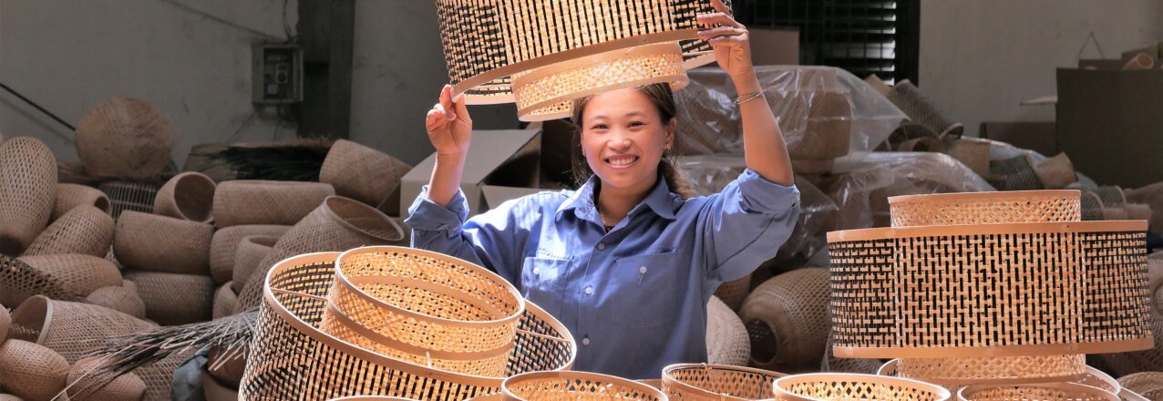 A woman working in a factory smiles for the camera while surrounded by woven bamboo baskets.